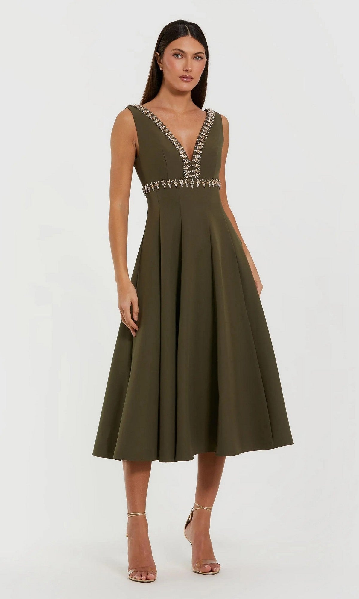 A woman models the olive Mac Duggal 21159 midi wedding guest dress featuring a sleeveless deep V-neckline and silver embellishments, set against a plain light background.